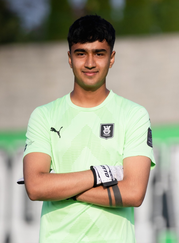 Ein junger Mann in einem grünen T-Shirt steht auf einem Fußballfeld und hat die Arme verschränkt, er strahlt, wahrscheinlich ein Torwart, mit einem unscharfen Hintergrund.