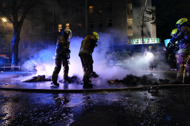 Feuerwehrleute in Helmen stehen nachts auf einer Straße mit Rauch, der aus einem Rohr aufsteigt, und halten Gegenstände, während Fahrzeuge und Gebäude mit beleuchteten Fenstern den Hintergrund bilden.