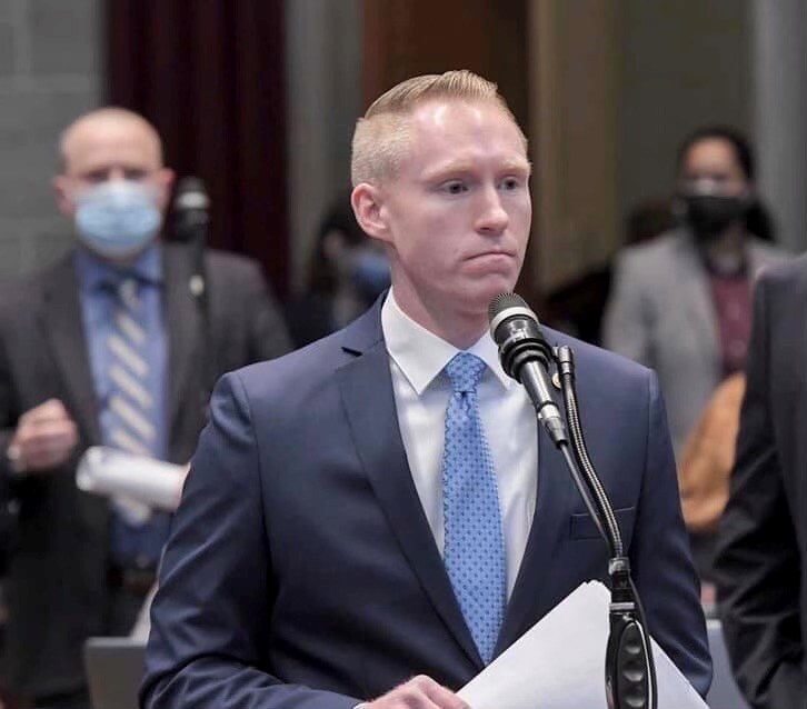 Ein Mann in Anzug und Krawatte spricht an einem Pult mit Papieren vor einem Mikrofon, während einige Menschen hinter ihm stehen und eine Vorhangwand im Hintergrund zu sehen ist.