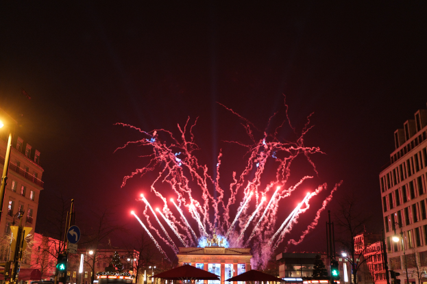 Eine belebte Straßen Szenerie in Berlin an Silvester, gefüllt mit Menschen, Fahrzeugen und Gebäuden, beleuchtet von Feuerwerk und Gebäudelichtern, schafft eine festliche Atmosphäre.