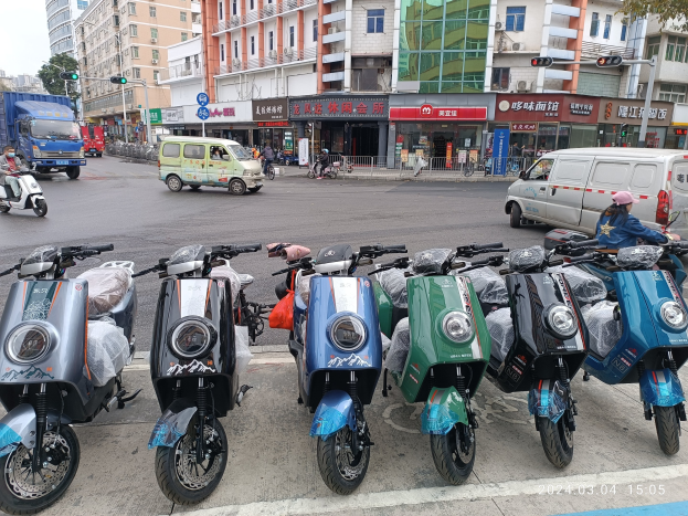 Eine Reihe von E-Scootern, die auf einer Straße geparkt sind, mit einer Person in einer Mütze davor, vor Gebäuden, Ampeln, Bäumen und einem klaren Himmel.