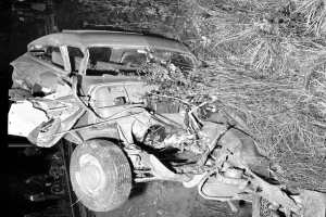 Ein Schwarz-Weiß- Foto eines schwer beschädigten Autos am Straßenrand mit einer zerquetschten Front, zersplitterten Fenstern, einem Baum im Hintergrund und verstreuten Gegenständen um die Unfallstelle.
