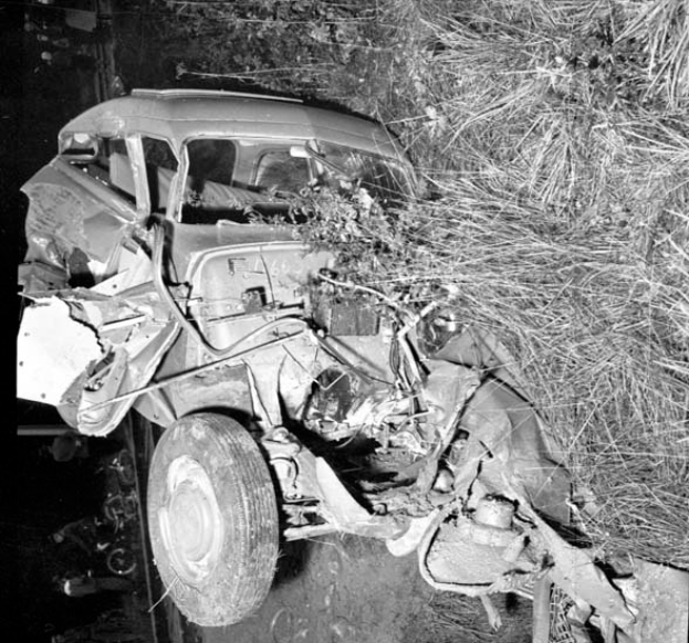 Ein Schwarz-Weiß- Foto eines schwer beschädigten Autos am Straßenrand mit einer zerquetschten Front, zersplitterten Fenstern, einem Baum im Hintergrund und verstreuten Gegenständen um die Unfallstelle.