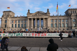 Eine Gruppe von Menschen hält ein Transparent mit der Aufschrift "Zukunft ist ein Mensch Neustar ima" vor dem Reichstaggebäude in Berlin, Deutschland, mit seinen architektonischen Details und umgeben von Flaggen unter einem bewölkten Himmel.