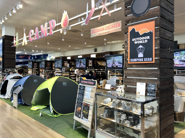 Ein Geschäft mit verschiedenen Campingausrüstungen, darunter Zelte, Infotafeln, eine Glasvitrine mit Gegenständen, Fernseher, Regale und andere Waren.
