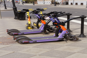 Eine Reihe von E-Scootern, die auf der Straßenseite geparkt sind, mit einem Mülleimer im Vordergrund und Gebäuden, Bäumen, Masten, Schildern und Fahrzeugen im Hintergrund.