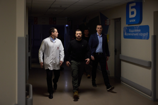 Eine Gruppe von Männern geht einen Krankenhausflur entlang und sieht besorgt aus, mit einer Tür auf der rechten Seite und Texttafeln auf der linken Seite.