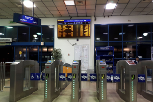 Ein Mann steht vor einer Metalldrehtür am Eingang eines Flughafens, mit Metalldetektoren, Informationsschildern, einem Topf mit einer Pflanze, einer Wandtafel, Deckenleuchten und einem Bildschirm im Hintergrund.