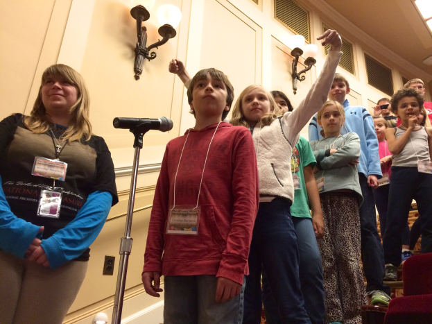 Gruppe von Kindern, die vor einem Mikrofon singen, mit einer Frau neben ihnen, Stühlen auf der rechten Seite und Wandlampen im Hintergrund.