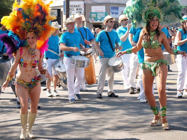 Zwei Mädchen tanzen mit Männern in blauen T-Shirts, weißer Farbe, Hüten und Schuhen, die Trommeln spielen, Geschäfte mit Plakaten und eine Stange im Hintergrund.