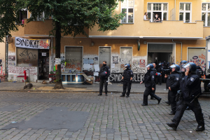 Eine Gruppe von Polizisten, einige mit Helmen und Waffen, geht eine Straße entlang vor einem Gebäude mit Fenstern, Läden und einer Fahne, mit einem Baum im Hintergrund.