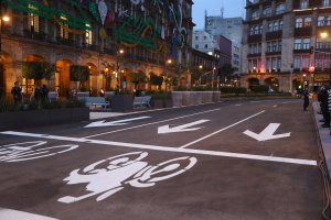 Stadtstraße mit einem ausgewiesenen Fahrradweg, umgeben von Gebäuden, Straßenmöbeln, Fußgängern, Grünflächen und einem klaren blauen Himmel.