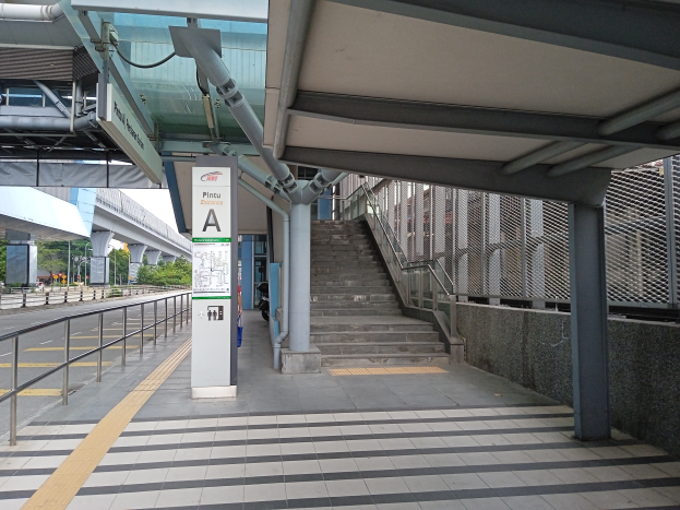 Ein Bahnsteig mit Treppe, eine Straße mit Geländer auf der linken Seite und ein Hintergrund mit Bäumen, Pfählen, Gebäuden und einer Brücke, mit Rohren und einer Tafel oben auf dem Bahnsteig.