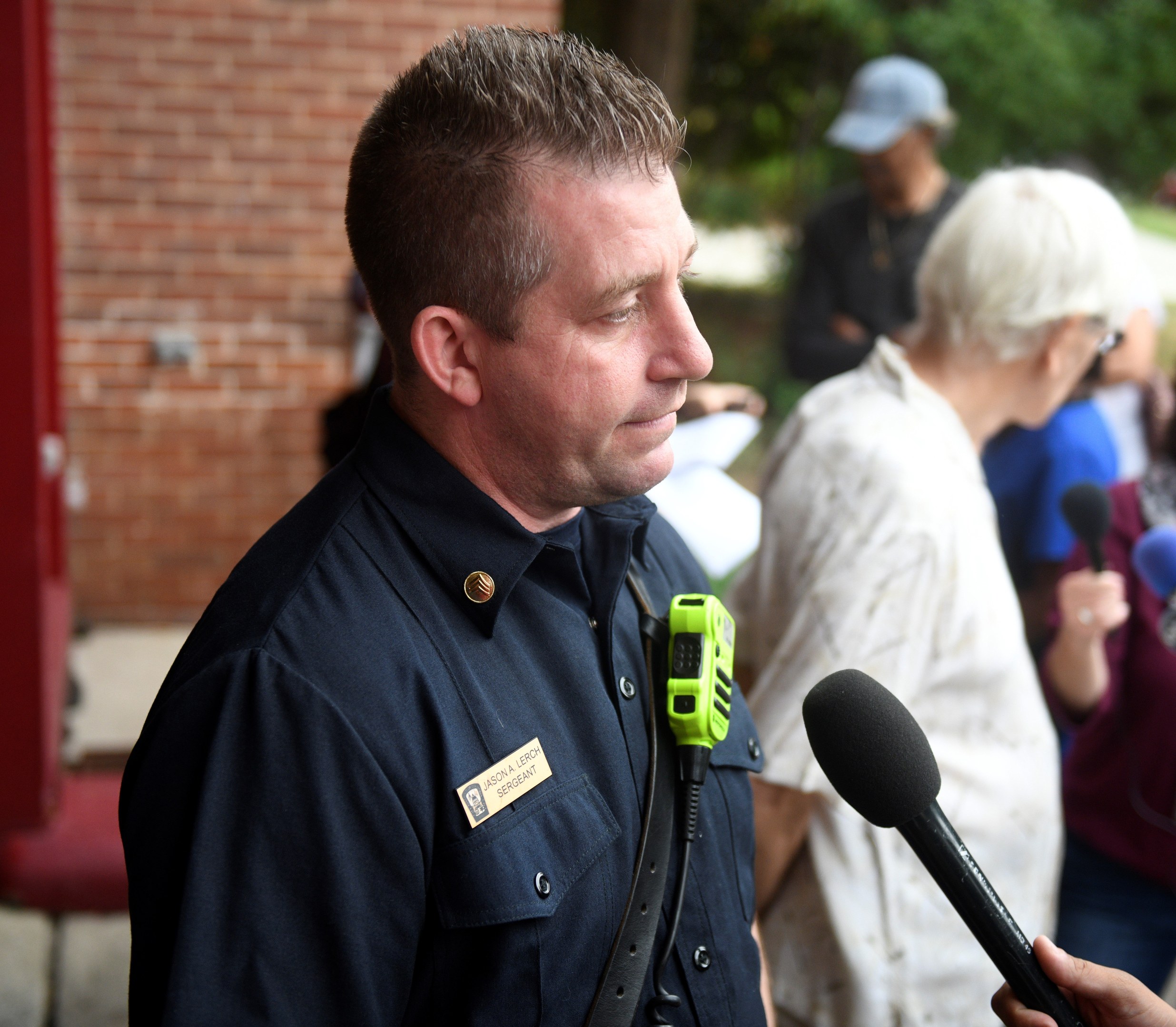 Ein Polizist in Uniform spricht mit Reportern, die Mikrofone halten, vor Bäumen und einer Backsteinwand.