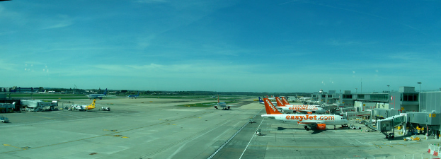 Eine Flughafen-Szene mit Flugzeugen, Gebäuden, Masten, Fahrzeugen und Bäumen unter einem Himmel mit einer Straße unten und einem Zaun im Vordergrund.