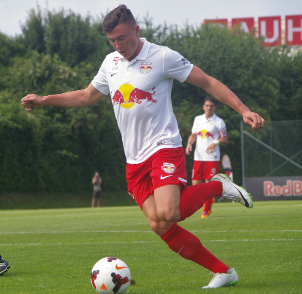 Ein professioneller Fußballspieler schießt den Ball auf einem Rasenfeld mit Bäumen und einem klaren blauen Himmel im Hintergrund, in der Nähe einer Tafel mit der Aufschrift "RB Leipzig."