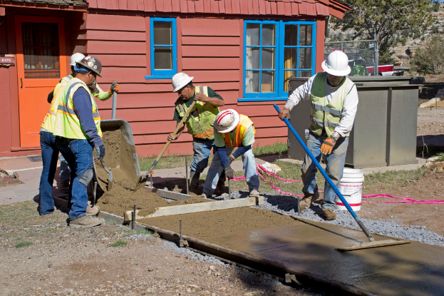 Eine Gruppe von Menschen, die draußen stehen und Werkzeuge halten, mit einer Betonmischung und einem Eimer in der Nähe, einem Gebäude, einem Zaun und einem Baum im Hintergrund, unter einem Himmel mit Gras zu ihren Füßen.