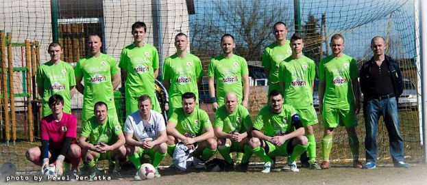 Gruppe von Männern in grünen T-Shirts auf einem Fußballfeld mit einem Ball davor, neben einem Torpfosten, mit Bäumen und Gebäuden im Hintergrund.