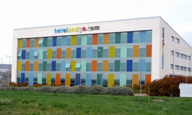 Großes modernes Hotelgebäude mit vielen Fenstern, umgeben von Grünflächen, Fahrzeugen und einem bewölkten Himmel, mit "hotel and go"-Schilder.