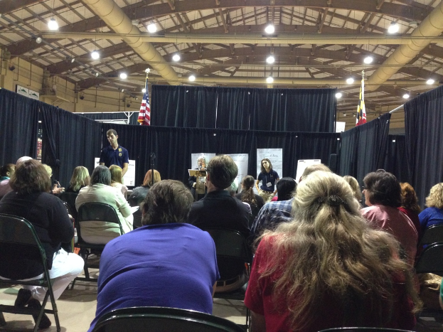 Eine Gruppe von Menschen, die auf Stühlen sitzen, mit einer Person, die an einem Rednerpult steht, in einem Raum mit Transparenten, blauen Vorhängen, Fahnen und Deckenleuchten.