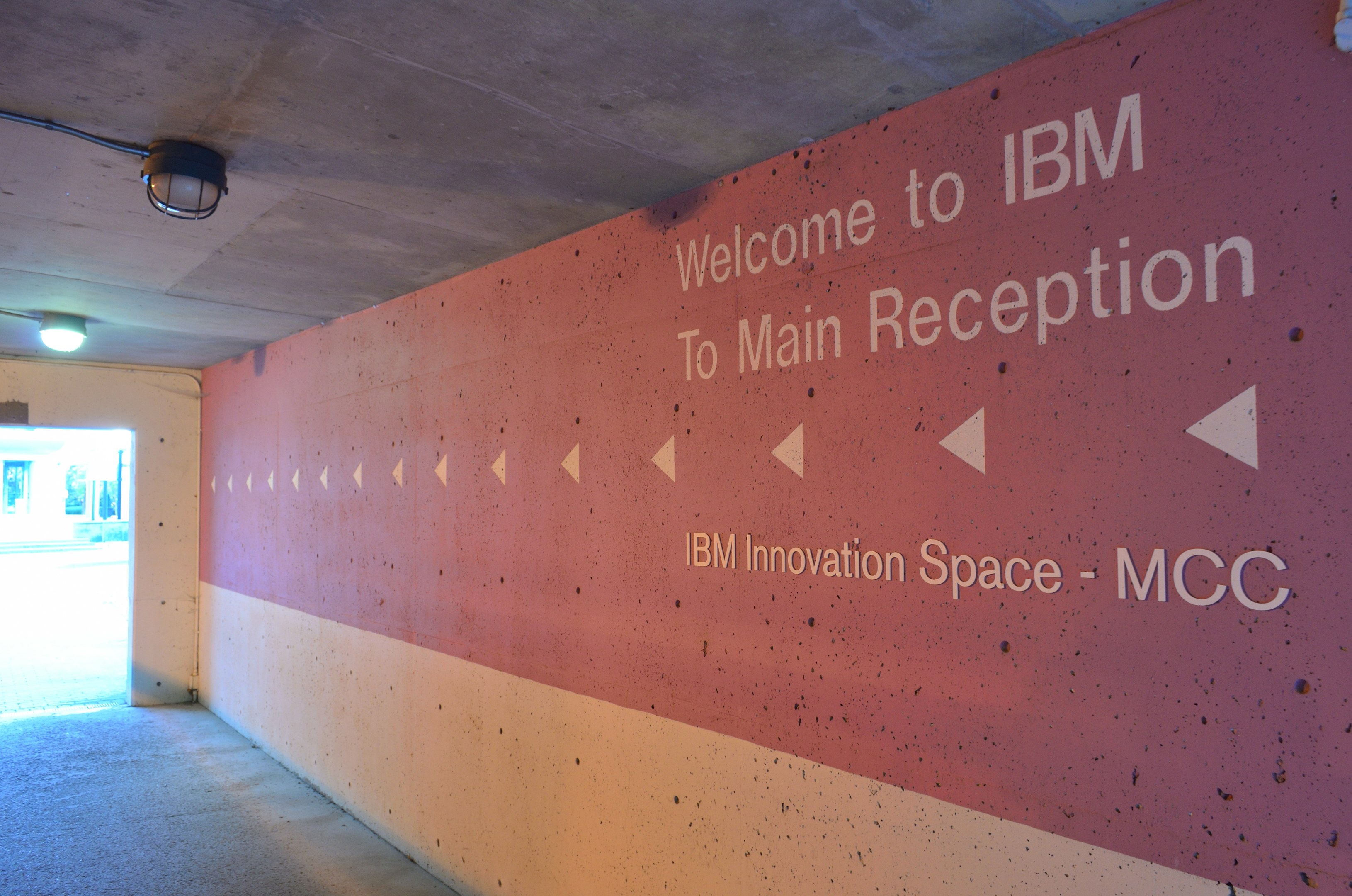 Eine Wand mit den Worten "Willkommen bei IBM" beleuchtet von Deckenlampen, mit einer Tür im Hintergrund beschriftet mit "IBM Innovation Space MCC."