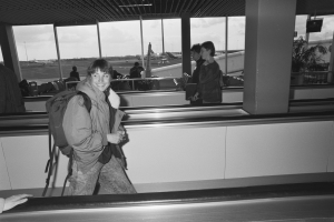 Eine Frau lächelt, während sie durch einen Flughafen geht und eine Tasche trägt, umgeben von Menschen, Flugzeugen, Pfählen, Bäumen und einem klaren blauen Himmel.