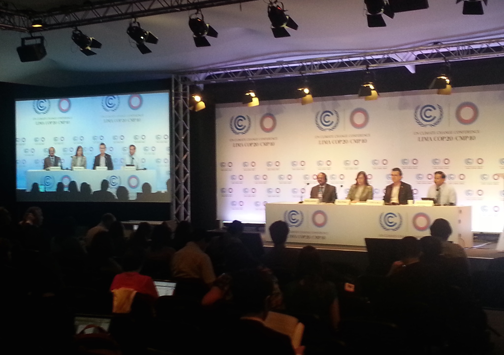 Menschen sitzen auf Stühlen auf einer Bühne mit Namensschildern und Mikrofonen auf einem Tisch, Zuschauer im Publikum, ein Projektionsschirm und Beleuchtung an Eisenstangen im Hintergrund.