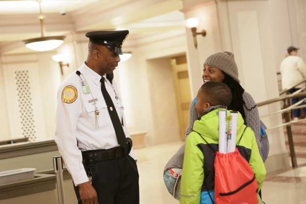 Ein Polizeibeamter spricht mit einer Frau in einer Mütze und einem Kind an einem Flughafen, mit anderen im Hintergrund.
