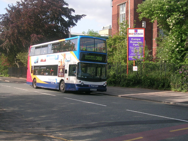 Ein Bus fährt auf einer Straße mit einem Metallzaun dahinter, einer Tafel in der Nähe und Bäumen, Gebäuden und Himmel im Hintergrund.
