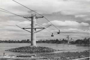 Schwarzes und weißes Foto einer Seilbahn, die eine von Pfählen getragene Stromleitung hochfährt, umgeben von Steinen, Gras, Holzstämmen, Wasser, Bäumen und einem bewölkten Himmel.