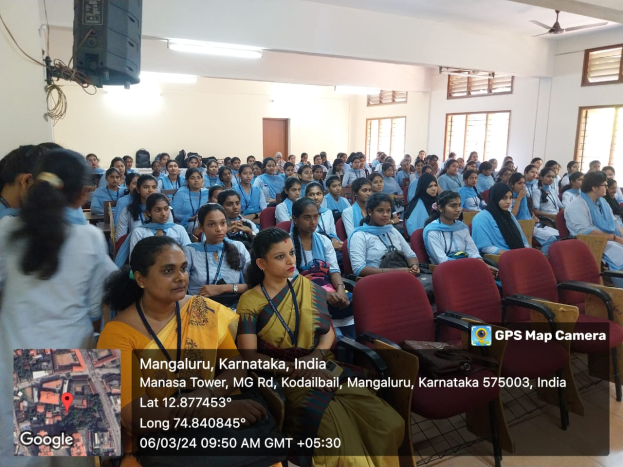 Eine Gruppe von Menschen, die in Stühlen in einem Raum mit einer Wand, Fenstern auf der rechten Seite, Deckenleuchten und einem Ventilator und einem Redner auf der linken Seite sitzen, mit Text und einer Karte unten.