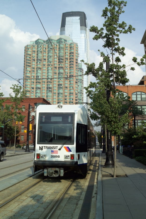 Ein Zug auf Schienen mit elektrischen Kabeln darauf, umgeben von Bäumen, Polen und Gebäuden, mit einem Wolkenkratzer und Himmel im Hintergrund.