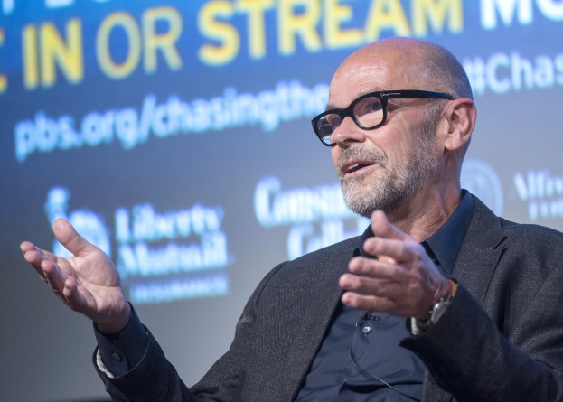 Steve Ballmer, Gründer und CEO von PBS, in einem Anzug und mit Brille, der in ein Mikrofon spricht, mit einem Bildschirm im Hintergrund, der Text anzeigt.