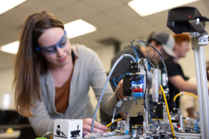 Eine Frau mit Schutzbrille arbeitet an einem Roboter in einem Labor, umgeben von anderen, mit Gegenständen auf dem Tisch und Deckenbeleuchtung.