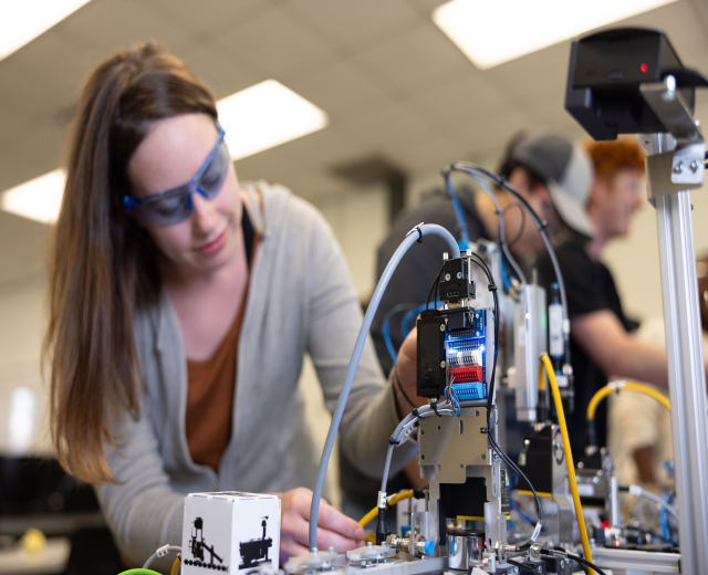 Eine Frau mit Schutzbrille arbeitet an einem Roboter in einem Labor, umgeben von anderen, mit Gegenständen auf dem Tisch und Deckenbeleuchtung.