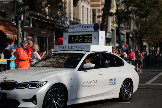 Ein weißer BMW mit einer Uhr auf dem Dach fährt eine Straße entlang, mit einer Person darin und einer Gruppe von Menschen, die auf der Straße stehen; Gebäude, Bäume und Ampeln sind im Hintergrund zu sehen.