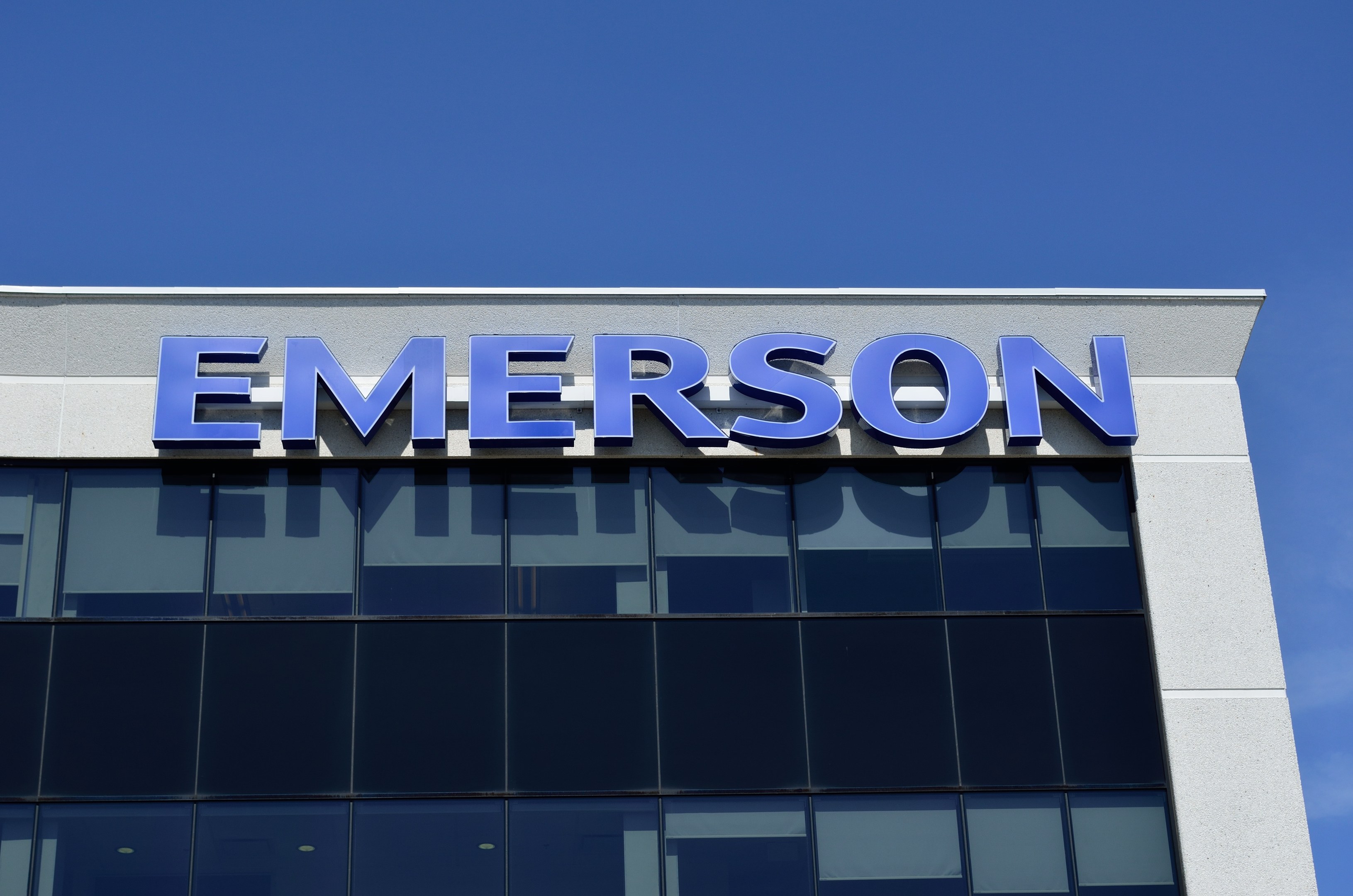 Emerson-Logo an einer Gebäudefassade mit dem Text "Emerson investiert 1,5 Milliarden US-Dollar" vor einem klaren blauen Himmel.