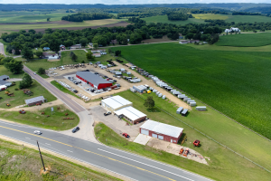 Luftbild einer ländlichen Farm mit Fahrzeugen, Scheunen, Bäumen, Gras, Pfählen und umliegenden Hügeln unter einem bewölkten Himmel.