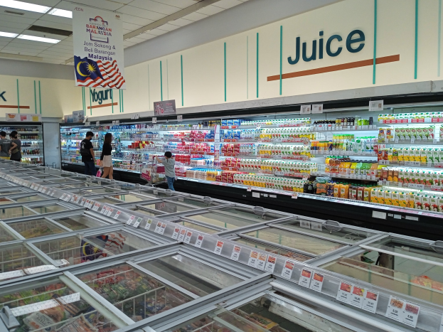 Ein gut sortierter Lebensmittelgang mit Regalen voller Getränke, Gemüse und anderen Lebensmitteln, einige Einkaufende und Deckenbeleuchtung.