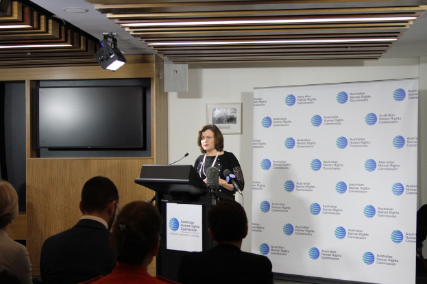 Eine Frau steht auf einer Bühne und spricht in ein Mikrofon, vor ihr sitzen Zuschauer, ein Banner an der Wand hinter ihr, ein Fernseher an der hölzernen Wand und Lampen an der Decke.