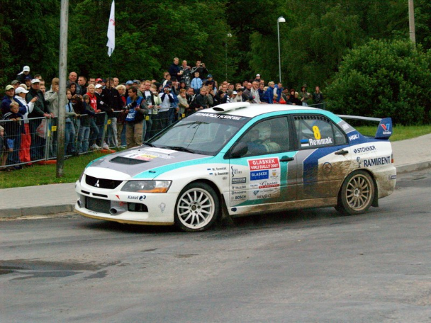 Ein Rallyeauto mit Text und Logos fährt an einer Menge vorbei, die Kameras hält, mit Absperrungen, Pfählen mit Fahnen und Bäumen im Hintergrund.