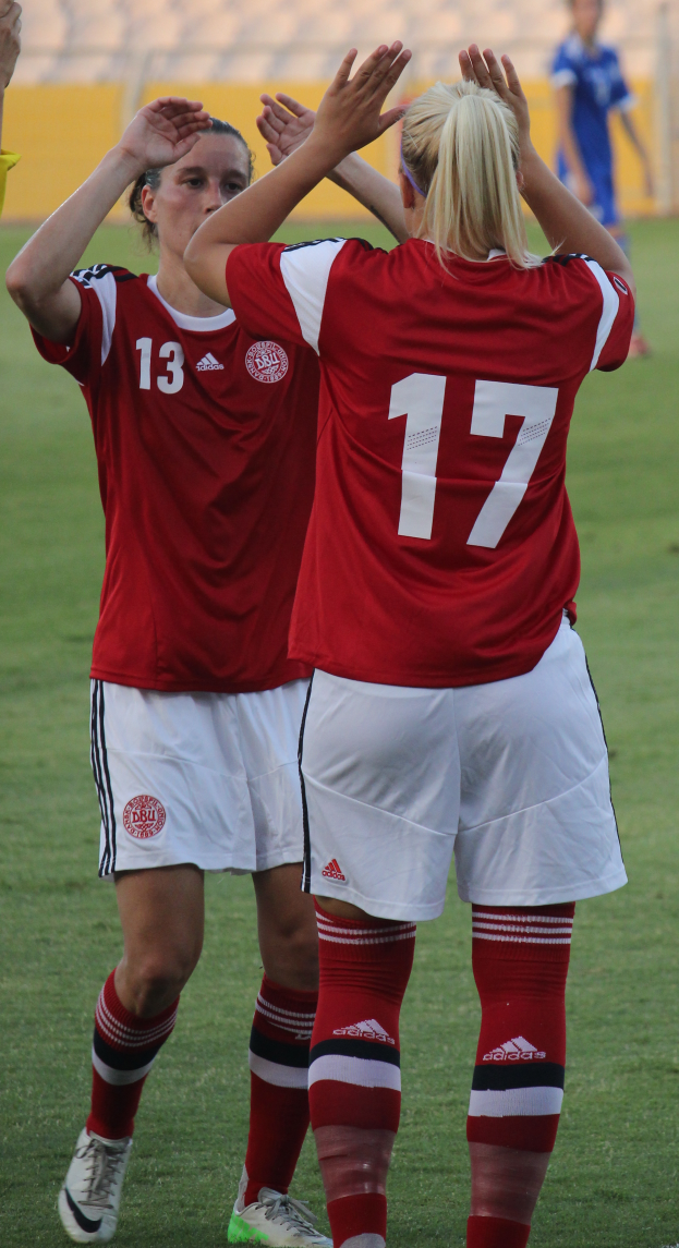 Zwei Frauen in roten T-Shirts und weißen Shorts stehen auf einem Fußballfeld und klatschen in die Hände, im Hintergrund eine Person in einem blauen Kleid, eine gelbe Tafel und ein paar Stühle.