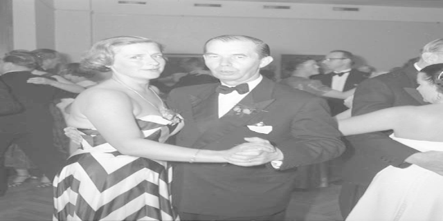 Ein Mann in einem schwarzen Blazer, weißem Hemd und schwarzen Hosen tanzt mit einer Frau in einem unbekannten Outfit auf einer formellen Veranstaltung und hält sich an den Händen, während sie zur Musik schaukeln.