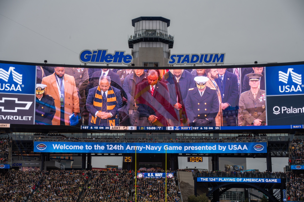 Eine große Menge Menschen in einem Stadion schaut ein Football-Spiel, mit einem zentralen Bildschirm, der Bilder und Text anzeigt, Schilder mit Text, Zuschauer in den Rängen, ein Tor, Bäume und einen klaren blauen Himmel im Hintergrund.