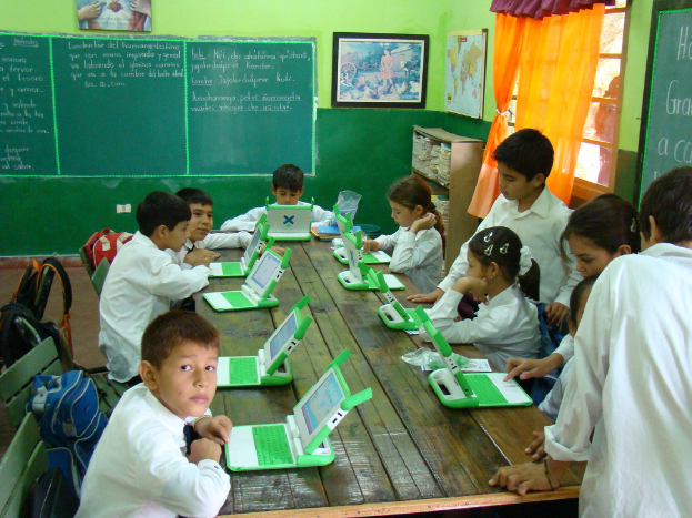 Kinder in Schuluniformen bedienen Computer in einem Klassenzimmer mit einer Tafel im Hintergrund.