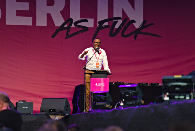 Ein Mann in einem weißen Hemd und braunen Hosen steht auf einer Bühne und hält ein Mikrofon in der Hand, hinter ihm ein pinkes Banner mit der Aufschrift "Berlin as Fuck" und Lautsprecher auf der Bühne.