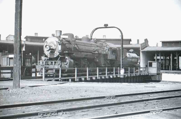 Ein Schwarz-Weiß-Foto eines Zuges auf Schienen mit Gebäuden und einem Mast im Hintergrund.