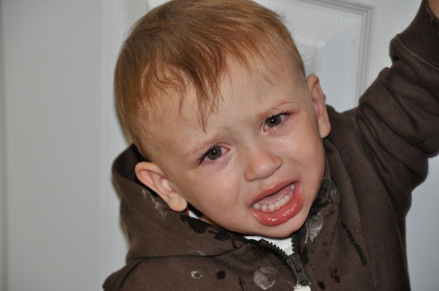Ein kleiner Junge in einer Jacke weint vor einer weißen Wand mit einer Tür im Hintergrund.