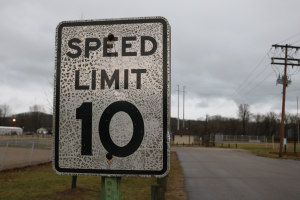 Ein Geschwindigkeitslimit-Schild am Rande einer Straße, umgeben von Gras, Strommästen mit Drähten, einem Zaun, Häusern, Bäumen und einem bewölkten Himmel im Hintergrund.