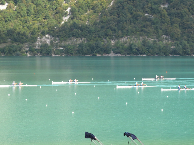Eine Gruppe von Menschen, die bei einem Wettbewerb auf einem Gewässer rudern, mit Bäumen und einem Hügel im Hintergrund und Kleidung auf Ständen am Ufer des Gewässers.